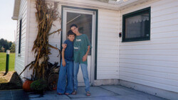Two people standing in a doorway of a white residential building with autumn decorations nearby.