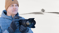 Photographer in winter gear holding a DSLR camera with hood lens in snowy landscape.