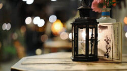 Black metal lantern with candle next to a book on wooden surface, with blurred bokeh lights in background.