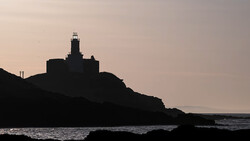 Lighthouse silhouetted against a twilight sky on a rocky coastal promontory.