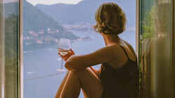 Woman sitting by a window overlooking a mountainous lake landscape, holding a wine glass.