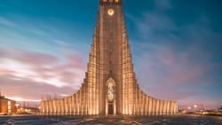 Illuminated modern church with soaring triangular architecture at dusk, featuring vertical ribbed columns and a glowing cross window.