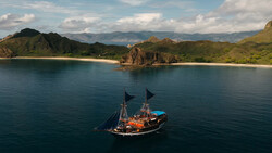 Traditional sailing vessel with dark hull and two masts anchored in calm turquoise water near a sandy beach and forested coastline.