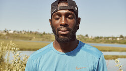 Man wearing light blue Nike t-shirt and dark cap standing outdoors with wetland landscape behind him.