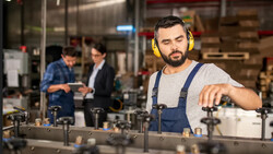 Factory worker in yellow hearing protection adjusts machinery while colleagues confer in the background.