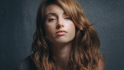 Woman with long wavy brown hair against a dark textured backdrop, looking downward with neutral expression.