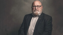 Headshot of a middle-aged man wearing glasses, dark blazer, and checkered shirt against neutral gray backdrop.