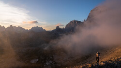 Hiker standing on a rocky slope with dramatic mountain peaks and misty valleys at golden hour.