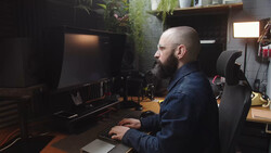 Bearded man working at a computer desk in a dark studio space surrounded by plants and equipment.