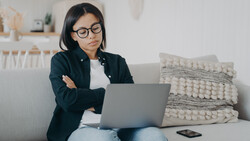 Woman wearing glasses and dark blazer sits on gray couch working on laptop with arms crossed.
