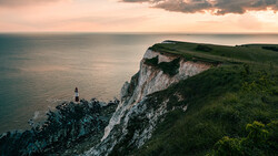 Lighthouse perched on white chalk cliffs overlooking calm sea at golden hour.