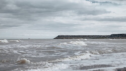 Rough ocean waves breaking on a beach under overcast sky with a rocky breakwater visible in the distance.