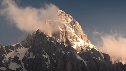 Snow-covered mountain peak with golden sunlit summit and dramatic clouds swirling around the ridges.