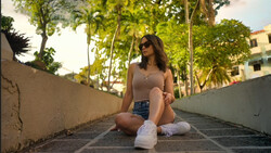 Woman sitting on a tree-lined pathway wearing sunglasses and casual clothing.
