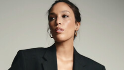 Woman in black pinstriped blazer against neutral background, looking upward with composed expression.