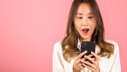 Woman in white shirt holding smartphone with surprised expression against pink background.