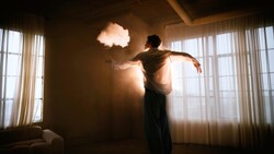 Person silhouetted against glowing cloud-shaped light suspended in a minimalist interior with large windows.