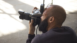 Photographer holding a camera with telephoto lens up to his eye, composing a shot in natural side lighting.