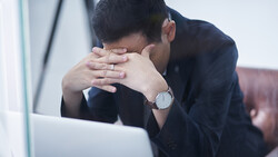 Man in dark blazer leaning forward with head in hands, facing laptop on desk.