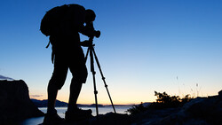 Silhouetted photographer on tripod against twilight sky and water.