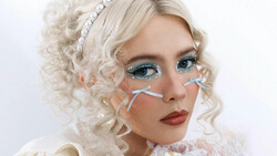 Portrait of a person with platinum blonde curly hair, dramatic eye makeup with blue eyeshadow, and decorative face accessories.