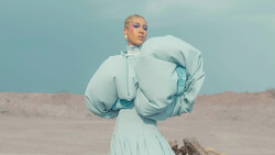 Person in flowing pale blue outfit with dramatic puffed sleeves standing in desert landscape against clear sky.
