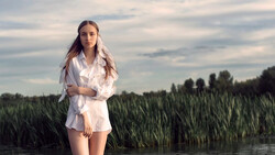 Woman in white shirt standing in shallow water beside tall green reeds at dusk.