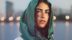 Woman wearing green headscarf looking at camera with urban waterfront and bokeh lights in background.