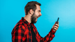 Bearded man in red plaid shirt holding smartphone against bright blue background.