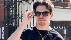 Man wearing sunglasses and black textured shirt with gold chain necklace adjusting his glasses.