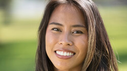 Woman with long dark hair smiling at camera outdoors with blurred green background.