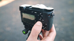 Hand holding a compact mirrorless camera with articulating screen and control dials visible.