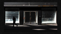 Silhouetted figure walking past storefront with illuminated window displays against dark facade.