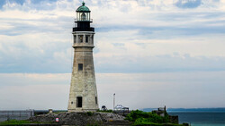 Stone lighthouse with green lantern room and gallery, standing on rocky coastal outcrop with calm ocean backdrop.