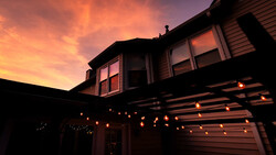 Two-story house silhouetted against vibrant orange and purple sunset sky, with warm string lights outlining the garage structure.