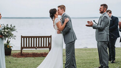 Groom and bride kiss during outdoor waterside ceremony as guests applaud.