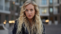 Woman with long blonde wavy hair looking at camera outdoors in urban setting.