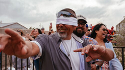 Man in white visor and sunglasses gestures toward camera while smiling at outdoor event with crowd in background.