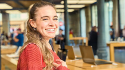 Young woman in red sweater smiling at camera in modern office space with wooden tables and blurred background activity.