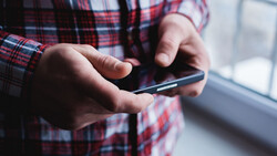 Close-up of hands holding a smartphone with a plaid shirt visible, shot near a window with natural lighting.
