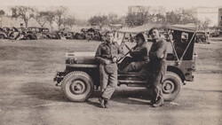 Three men in military uniforms standing beside a utility vehicle in an open field with vehicles and structures visible in the background.