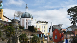 Hillside coastal town with white-domed chapel, yellow building, and colorful historic structures under partly cloudy sky.