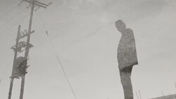 A tall, textured human figure stands against a utility pole in an overcast landscape.