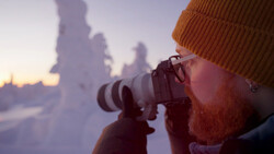 Bearded photographer with orange beanie holding telephoto lens to eye during golden hour, snow-covered landscape blurred in background.