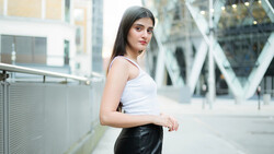 Young woman in white tank top and black leather skirt posing on urban pedestrian bridge.