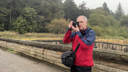 Male photographer in red and blue jacket holding a camera to his face while standing on a wooden boardwalk.