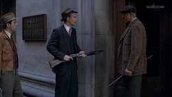 Three men in period clothing stand outside a post office; the central figure holds a rifle.