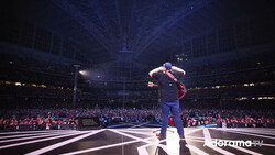 Performer on stage in large arena with dramatic blue overhead lighting and crowded audience.