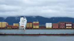 Large cargo ship stacked with colorful shipping containers against a mountainous coastline.
