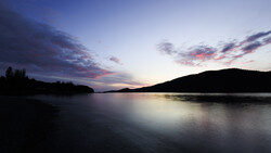 Twilight landscape with calm lake, forested shoreline, and mountains silhouetted against purple and pink sky.